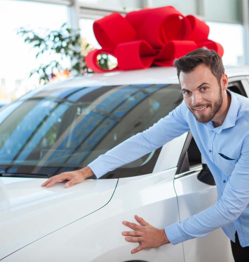Ein junger Mann streicht über die Kühlerhaube eines neuen Autos. Auf dem Autodach eine große rote Schleife.