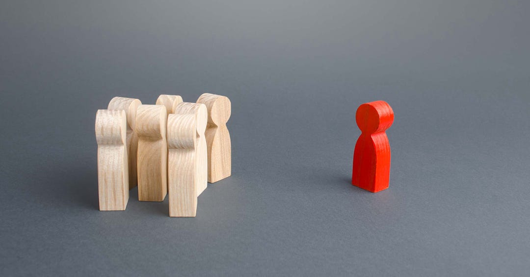 Einer Gruppe von hölzernen Spielfiguren steht eine rote Spielfigur gegenüber